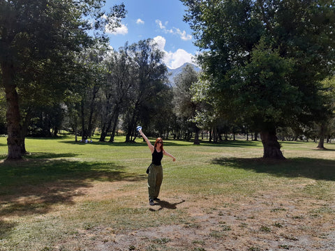 Une femme se tient au centre d'une plaine, avec des arbres au fond et deux grands de chaque côté. Elle est joyeuse et tend ses mains vers le ciel en signe de victoire.