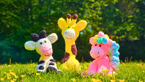 A group of stuffed toys sit together in the grass on a summers day, from left to right sit a knitted cow, giraffe and pink unicorn