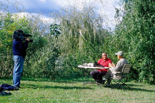 Ken being interviewed and filmed, sat in a green space next to a lake.