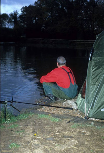 Ken wearing his fishing gear, looking out of on a fishing lake at dusk, next to a green tent.