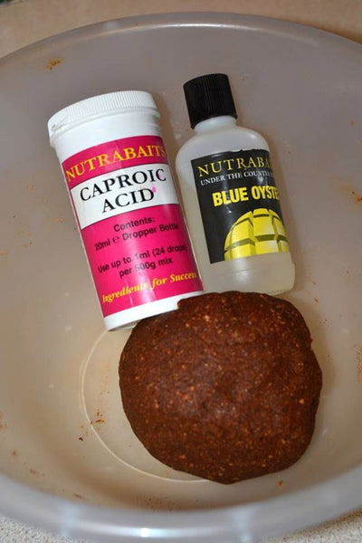 Photograph of a container with caproic acid and a second container with blue oyster attractor in front of a red round boilie for fishing