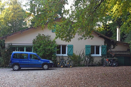 Chalet bungalow with gravel front, overhanging trees and a blue vehicle parked outside.