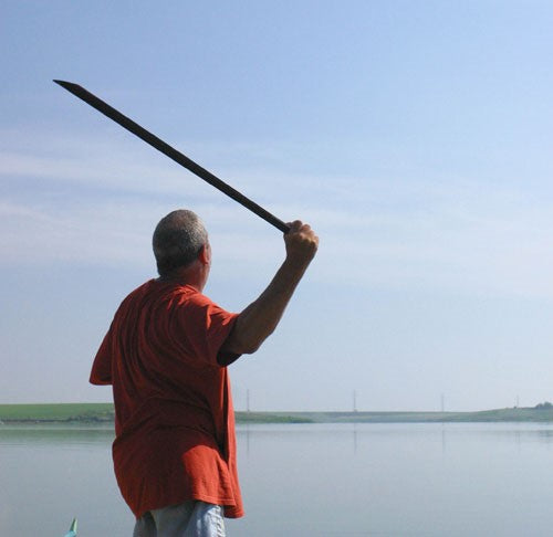 Ken holding a long throwing stick, in front of water.