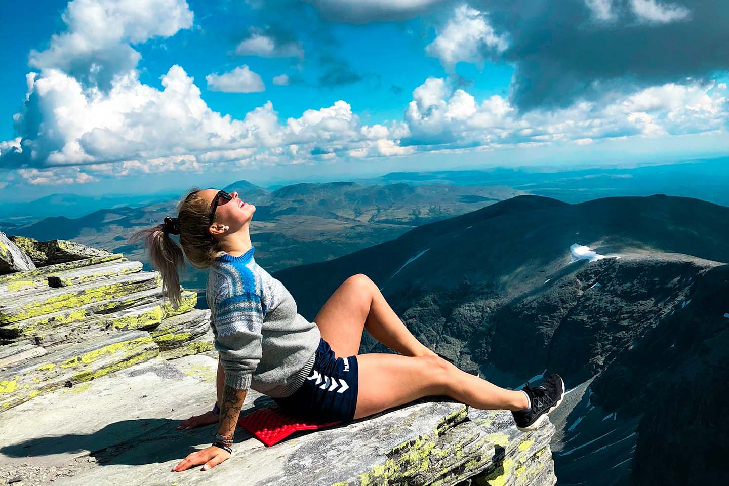 a woman happily sitting on peak of a mountain while looking up at the blue sky with clouds