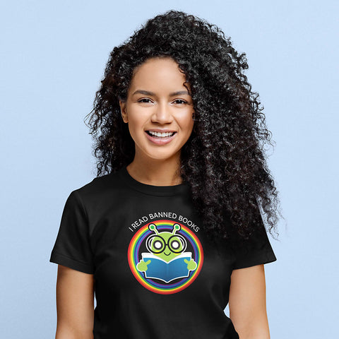 A woman of color wearing a black unisex t-shirt with an illustration of a bookworm holding up its middle fingers while reading a book inside a rainbow circle. The text says, “I read banned books” arched above the illustration.