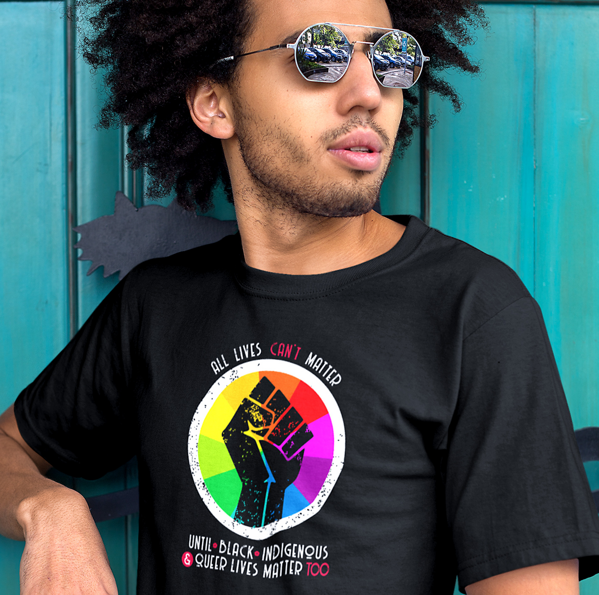 A Black man wearing a team purple tee with a graphic on the front of a circle with radial rainbow colors in it and a black fist over the colors. The text above the graphic is arced and reads “All lives can’t matter” and then horizontally below the graphic reads “until Black, Indigenous, & Queer lives matter, too”.