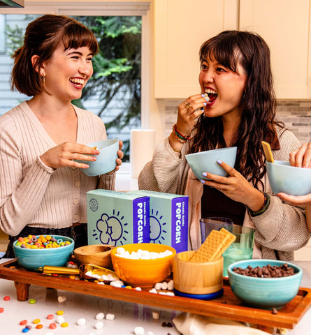 Women eating bowls of popcorn with a spread of toppings and boxes of kettle corn in front of them