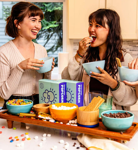 two women behind a popcorn bar that includes graham crackers, marshmallows, and chocolate chips