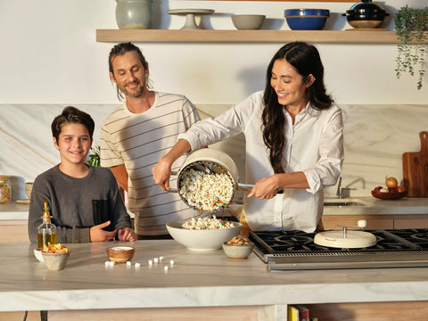 A woman pours popcorn from the Popper to a bowl while a man and young boy smile