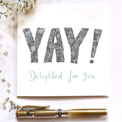 Product Image for celebration card that says YAY! Delighted for you, on the front. The word YAY! is illustrated in black and white intricate leafy detail. Delighted for you is hand written in a sage green underneath. Around the greeting card are some flowers and a gold pen for decoration.