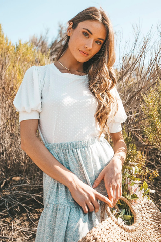 A woman in a flattering white blouse and high-waisted skirt stands outside. 