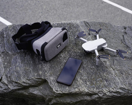 A set of drone flying equipment laid out on a rock surface. On the left is a DroneMask, used for first-person viewing from the drone's camera. To the right of the goggles is DJI mini drone. Beside the drone is a smartphone, used to view the camera feed inside the DroneMask.