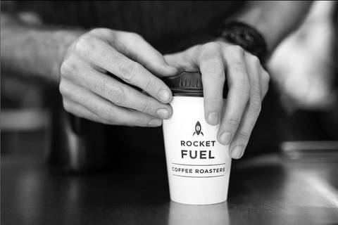 Black and white image of a  Rocketfuel take away cup with black logo printed on it with lid being places on
