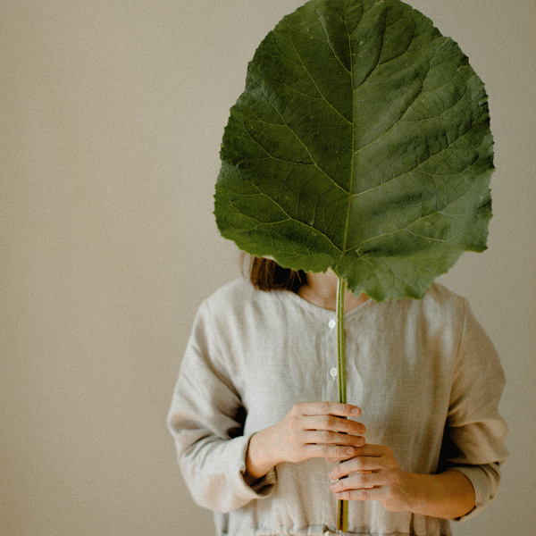 a person in a cream linen dress holding a giant plant leaf in front of face