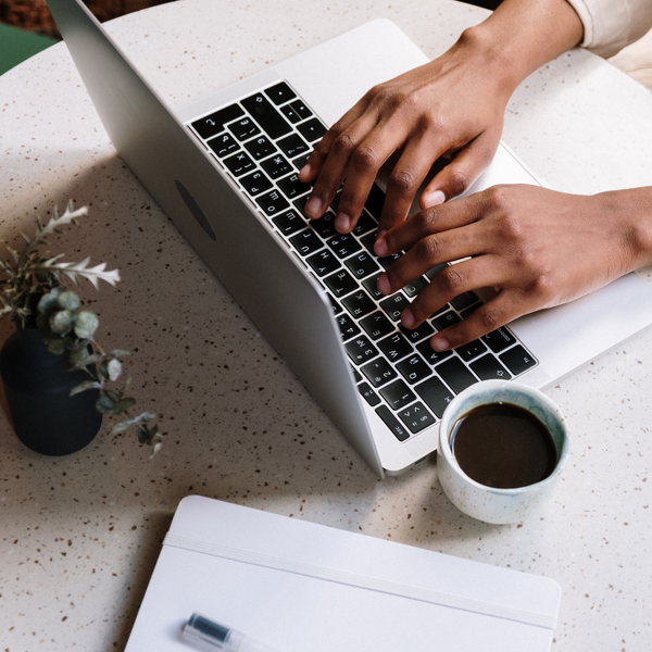 A person's hands from above, resting on a laptop keypad. On the table in a cup of black coffee and a small plant