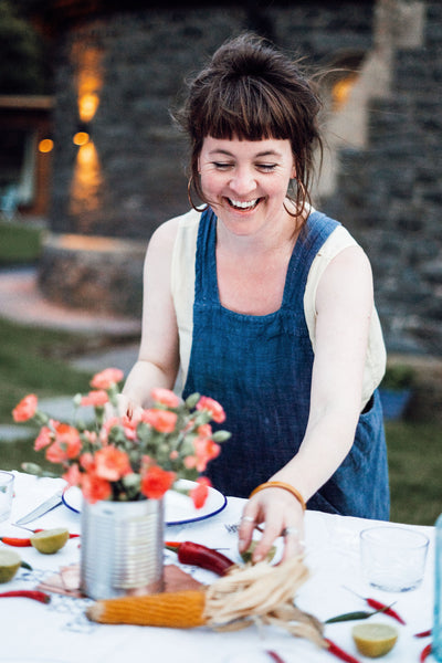 Java Bere laughs and attends to beautiful table display, with bright flowers in centre. She wears a denim blue pinafore dress, white t-shirt and large gold hoop earrings.