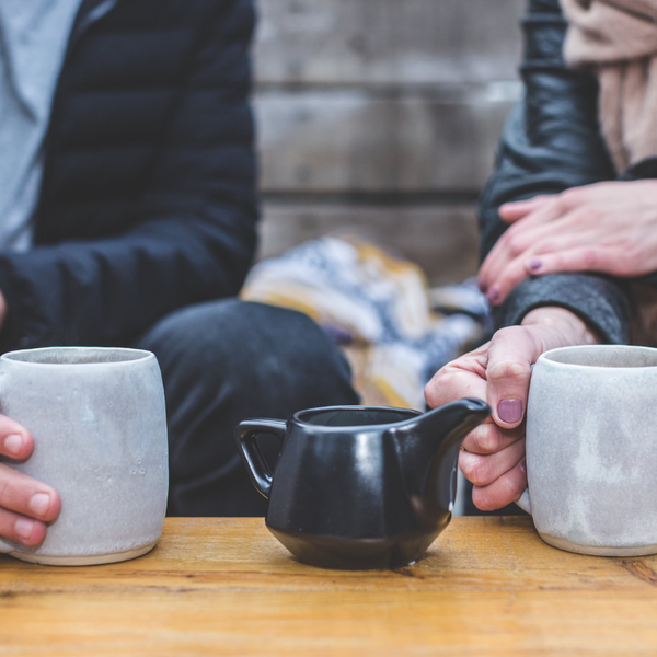 close up of two people holding warm drinks, with teapot on table beside them
