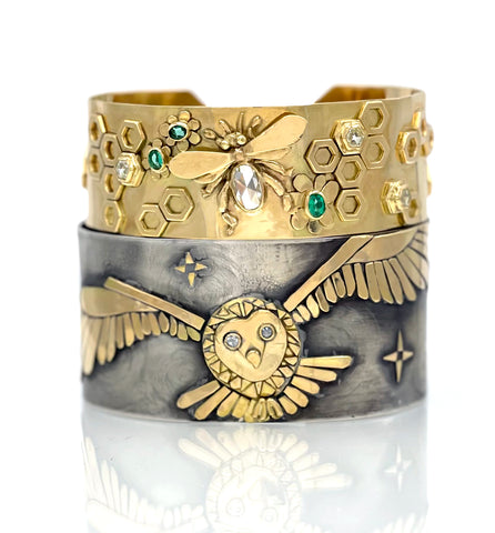 two wide cuff bracelets stacked one in solid gold with a bumble bee and one in sterling silver and solid gold with an owl