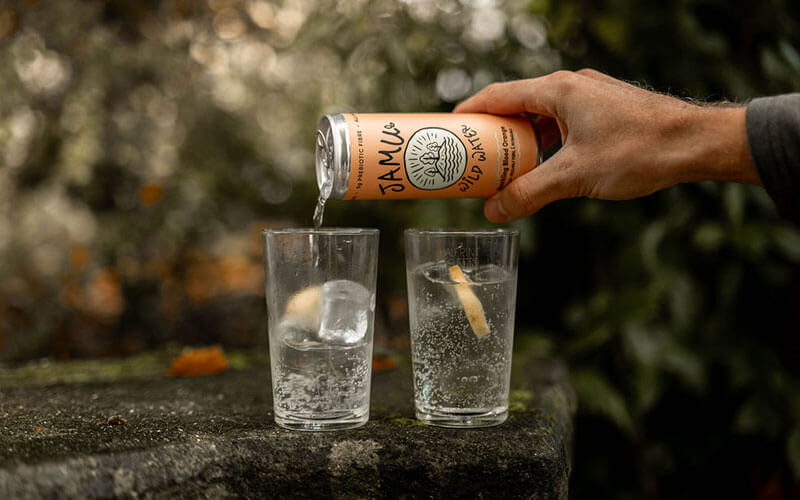 An orange coloured can of Blood Orange Jamu Wild Water being poured by a male hand into a glass with ice and a slice of lemon. An out of focus, wooded/garden environment is behind.
