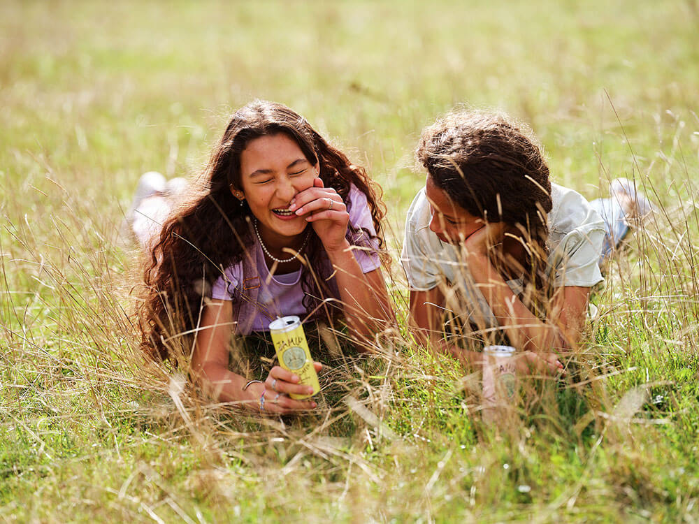 Two young girls laughing. They're lying in long grass and both drinking a can of Jamu.