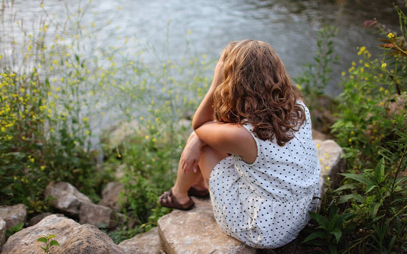 A girl in a white polka dot dress sits on a rock over looking still water. She leans her forehead against her left palm as though concerned.