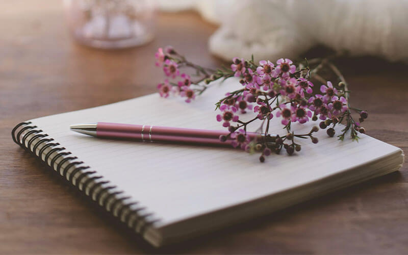 A ring bound journal folded open on a blank page. A pink pen and purple flowers sit on the page.