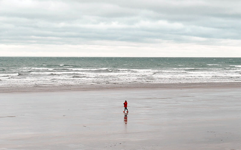 Looking out to sea, a person in a red coat with red shoes walks along an empty beach in North Devon. It's cloudy, with plenty of waves rollin in.