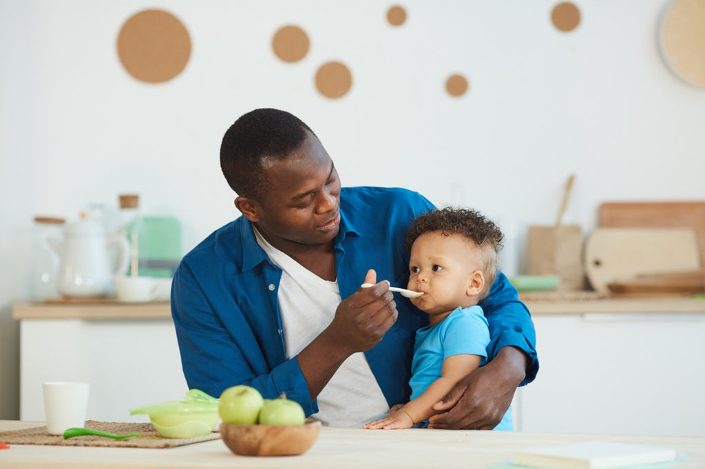 Portrait of happy dad feeding cute little boy sitting at kitchen table with green apples, copy space