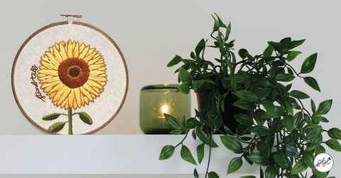 Sunflower embroidery hoop displayed on a shelf next to plant and candle.