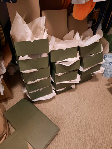 Three stacks of of green bakery boxes that are folded and stuffed with white tissue paper. The boxes are surrounded by clothes hanging above them since the scene is set in a clset.