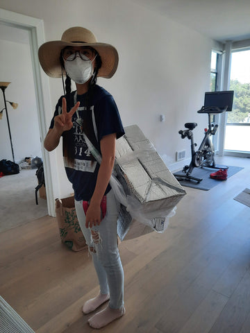 Girl with hat and mask on with a box tied to her back using plastic bags. The girl is holding up a peace sign.