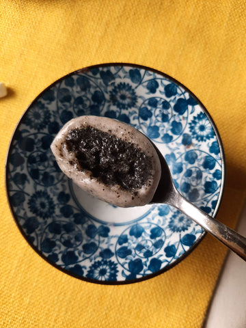 Cross section of black sesame tangyuan on a silver spoon. Blue and white patterned bowl in the background on top of a yellow table runner.