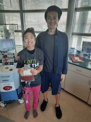 Annie and Jeffrey standing in a hospital clinic. Annie is holding up a bag of bone marrow donation she has just donated to Be the Match.