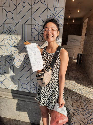 Annie holding up a white bag filled with churros in front of a white tiled wall with blue lines.