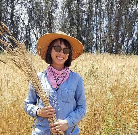 Farmer Mai holding a bundle of wheat smiling. A golden wheat field in the background.