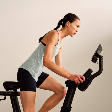Image of an woman exercising using the Carol Bike. She has long hair and pony tail. She's wearing a light blue top and dark shorts. She looks focused, eye on the ball!