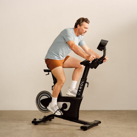Image of a man exercising with the CAROL bike. He's wearing a light blue t-shirt, light brown shorts, white socks and white and light grey trainers. He has longish curly hair and a 5-day beard. He is holding the bar with both hands and looking at the screen.