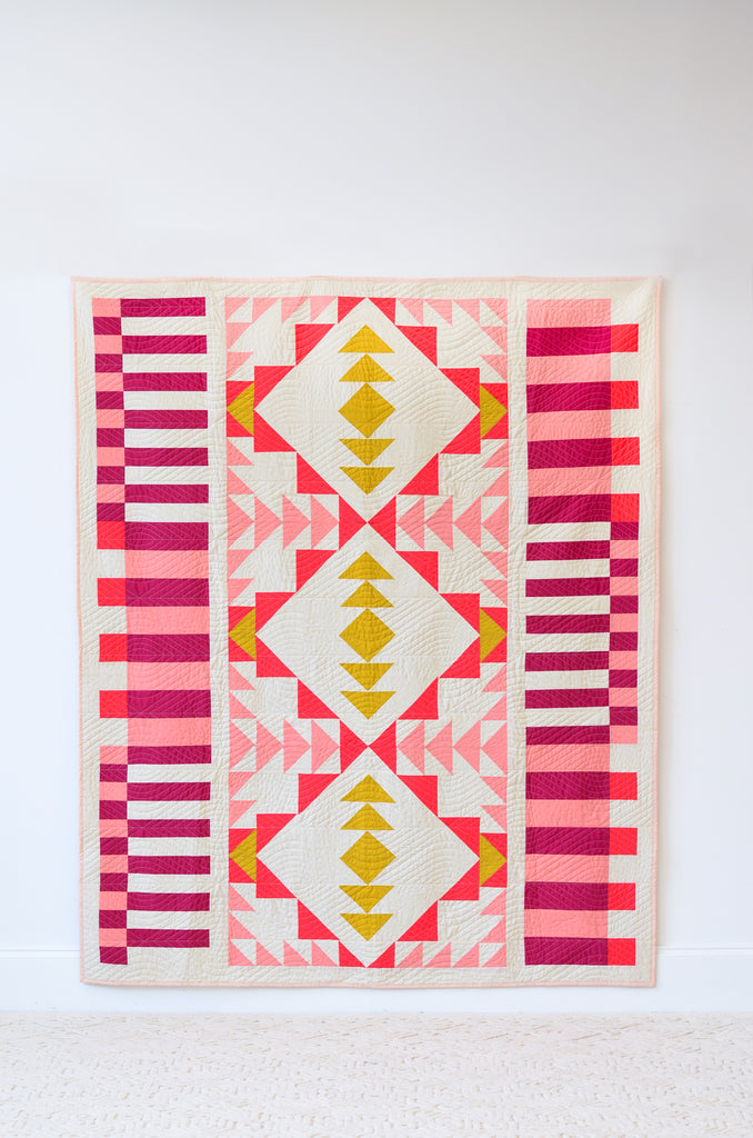 Open road quilt by toad and sew, red pink, and yellow quilt hanging on white wall 