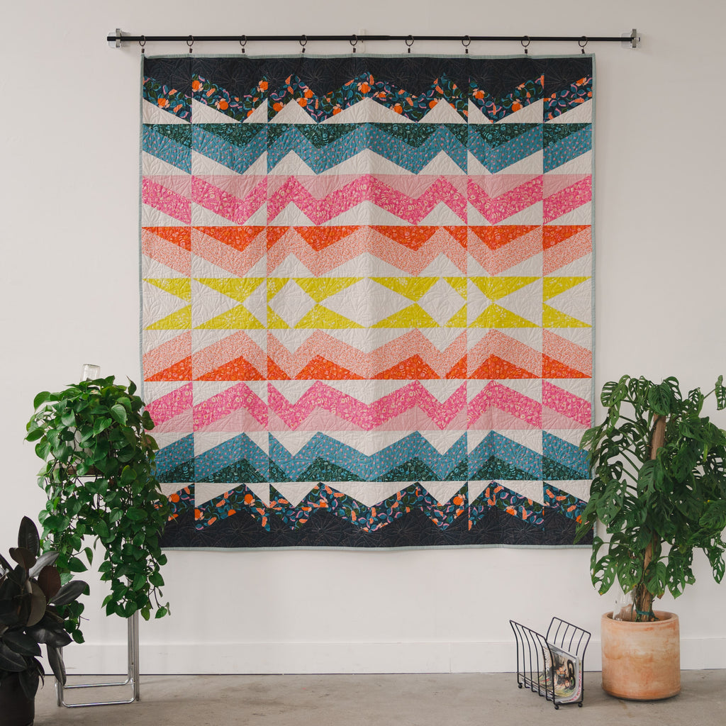 Quilt hanging on white wall with yellow and pink triangles that are radiating outward