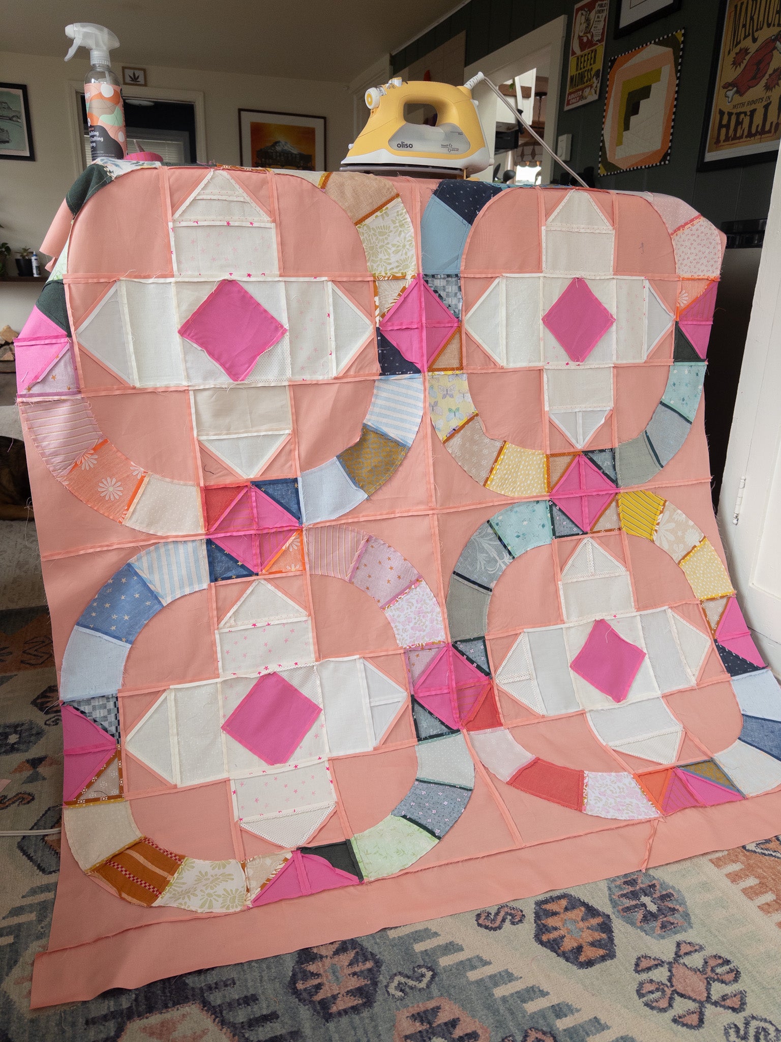 Nearly assembled scrappy Moonbeam baby quilt top draped over an ironing board in a cozy living room: four peach-background circular blocks with colorful scrap-pieced arcs and hot pink diamond centers, seams pressed, with an iron and starch spray visible on top.