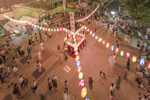 View of the square in front of the Nippori train station decorated for the Obon festival with a yagura tower illuminated with paper lanterns where a girl in traditional costume is playing taiko drum.