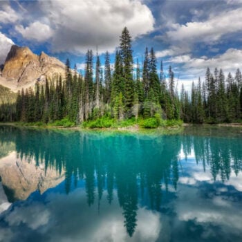 emerald colored lake with tall pines and mountains in distance