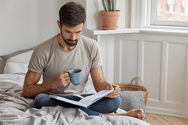 self-care-male-reads-book-during-day-off-involved-reading-drinks-hot-beverage-sits-crossed-legs-bed-wears-casual-t-shirt-trousers-people-knowledge-education-leisure