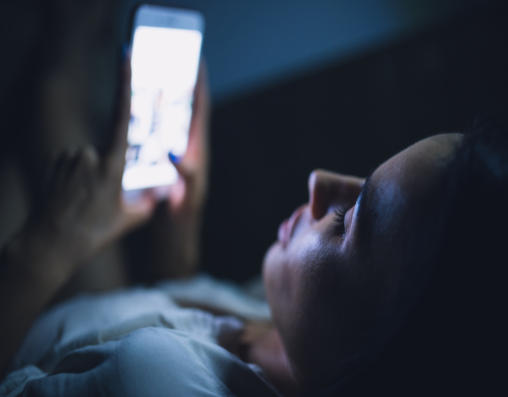 Blue light should be avoided in the evening and at night before going to bed. The picture shows a woman's face that is really close to the phone and a blue light is coming from the screen. He is just about to go to sleep, but the blue light does not promote falling asleep or sleep quality. Blue light inhibits melatonin production.