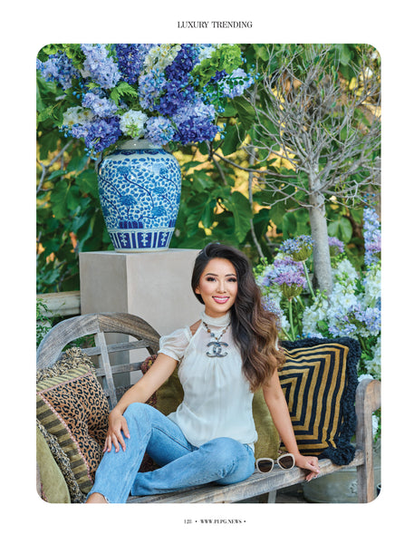 Victoria Tai of Winward Home sits on bench in front of large faux delphinium floral arrangement