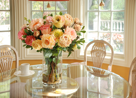 Faux flower arrangement in a clear glass vase sits in the center of an elegant glass-top table near a sun-filled bay window.