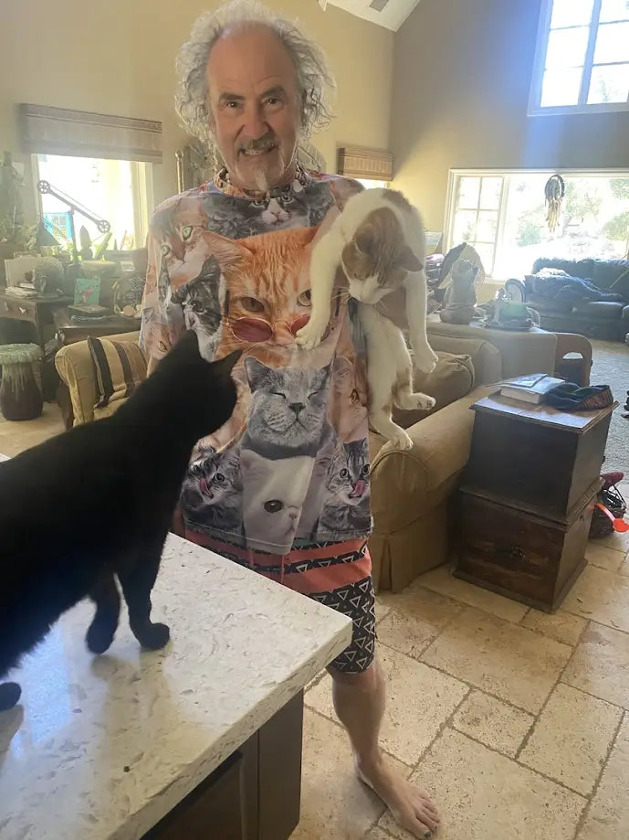 Smiling man in a cat-themed shirt holding a cat, with another cat on a counter.