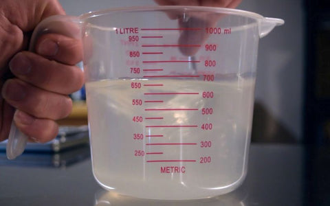 A hand mixes a solution of distilled water and light malt extract in a clear container.