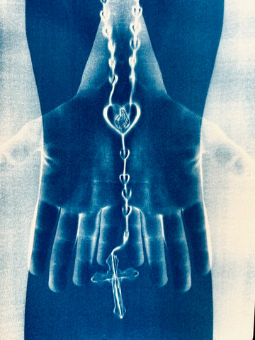 Image is a cyanotype print in deep blue tones of a pair of hands holding a rosary