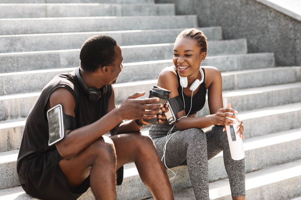 Fit Black Couple Taking Rest After Workout Outdoors, Sitting On Urban Stairs, Drinking Water And Coffee And Chatting, Free Space